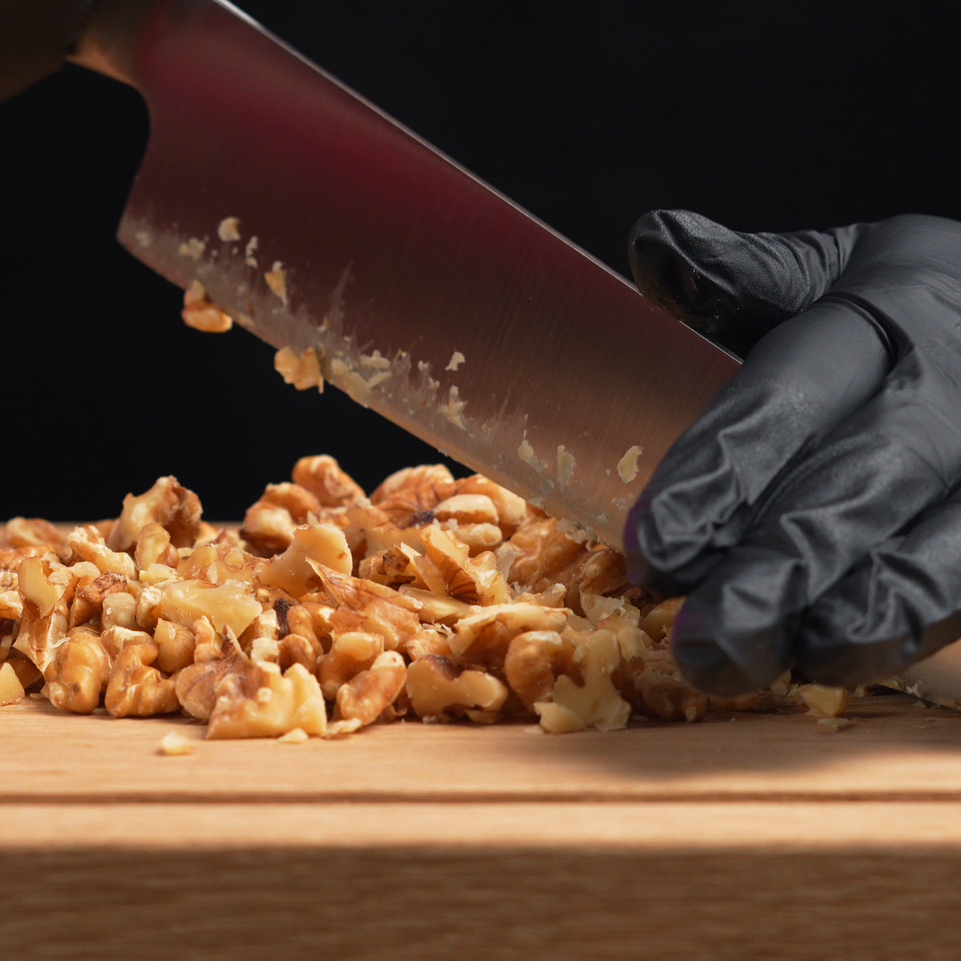 Person wearing black gloves chopping walnuts on a wooden cutting board with a knife.