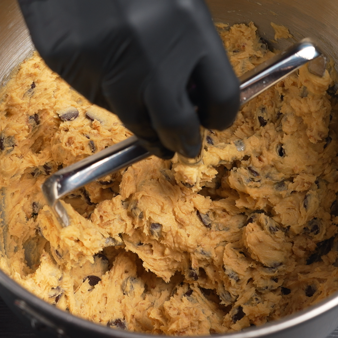 Cookie dough with chocolate chips being mixed in a metal bowl with a dough mixer.