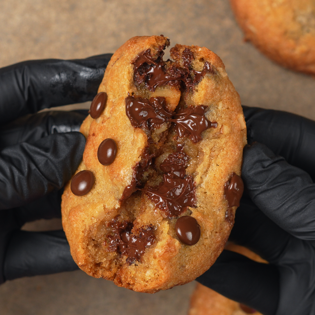 Chocolate chip cookie held by a person wearing black gloves on a brown surface.