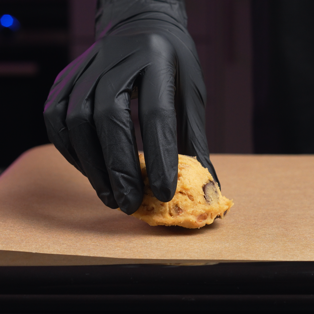 Hand wearing a black glove holding a cookie dough ball on a baking sheet.