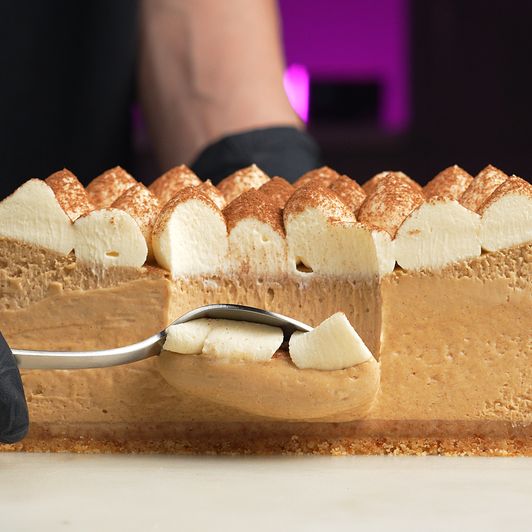 A close-up of a layered dessert cake with a golden-brown mousse filling, cream-colored sponge layers, and piped meringue peaks on top, with a spoon lifting a slice in a kitchen setting.
