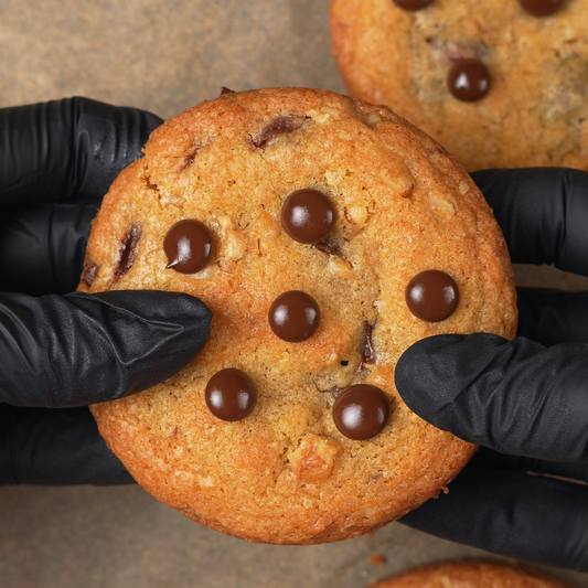 Chocolate chip cookies held by black-gloved hands on a brown surface