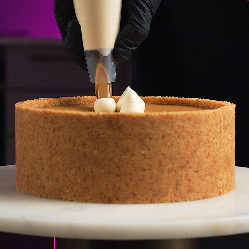 A close-up of a person wearing black gloves piping cream onto a round cake with a golden-brown crumb crust, shown on a white turntable.