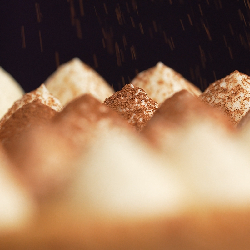 Close-up of a meringue-topped pastry or cake with piped peaks dusted in cocoa powder, set against a dark background with a purple light and falling sprinkle effect.