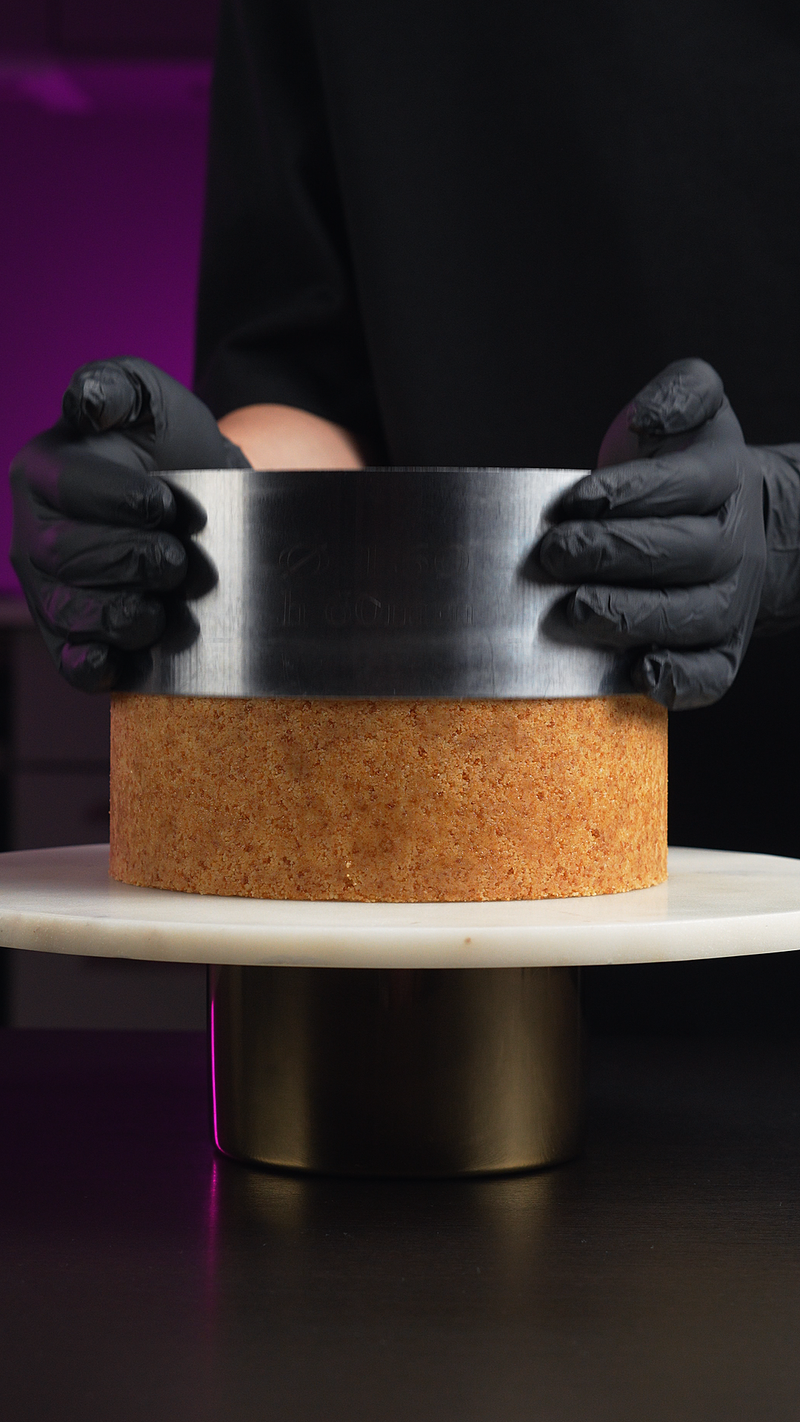 A person wearing black gloves holds a round metal ring mold over a square wooden cake or pastry base on a white round turntable.