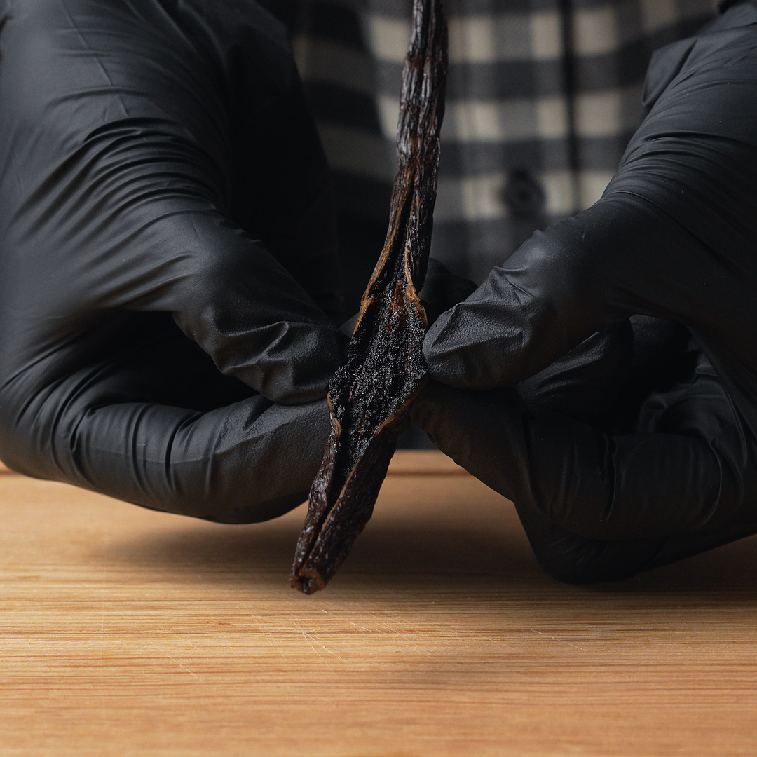 Person wearing black gloves holding a vanilla pot on a wooden surface